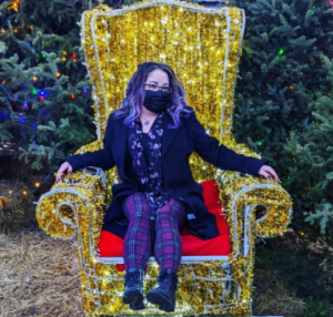 Author seated on a large chair made of twinkling lights, wearing purple plaid pants, black boots, and a long black coat.